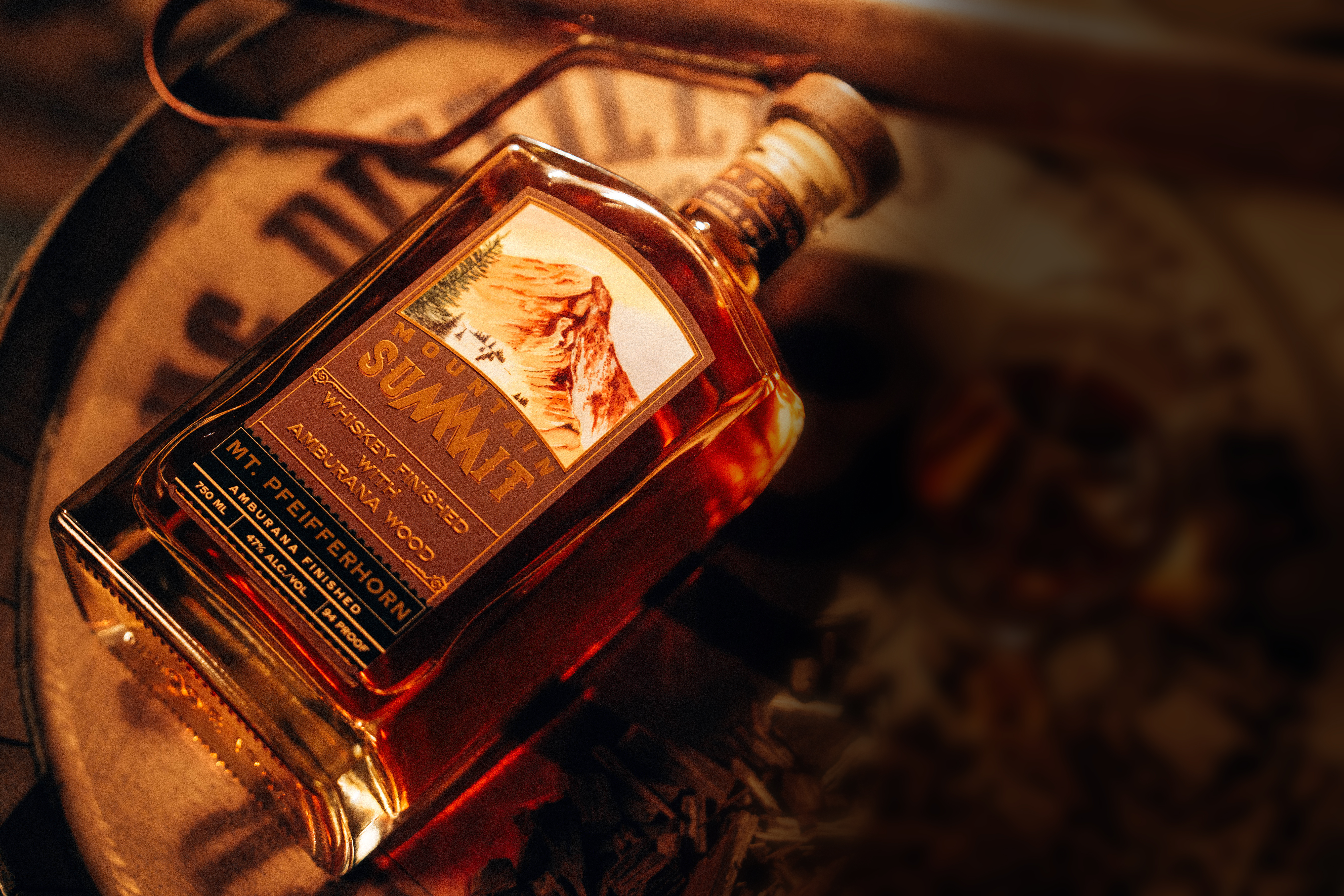 Mountain Summit Whiskey Bottle - Professionally photographed. Sitting atop an aging barrel with a glass next to it.